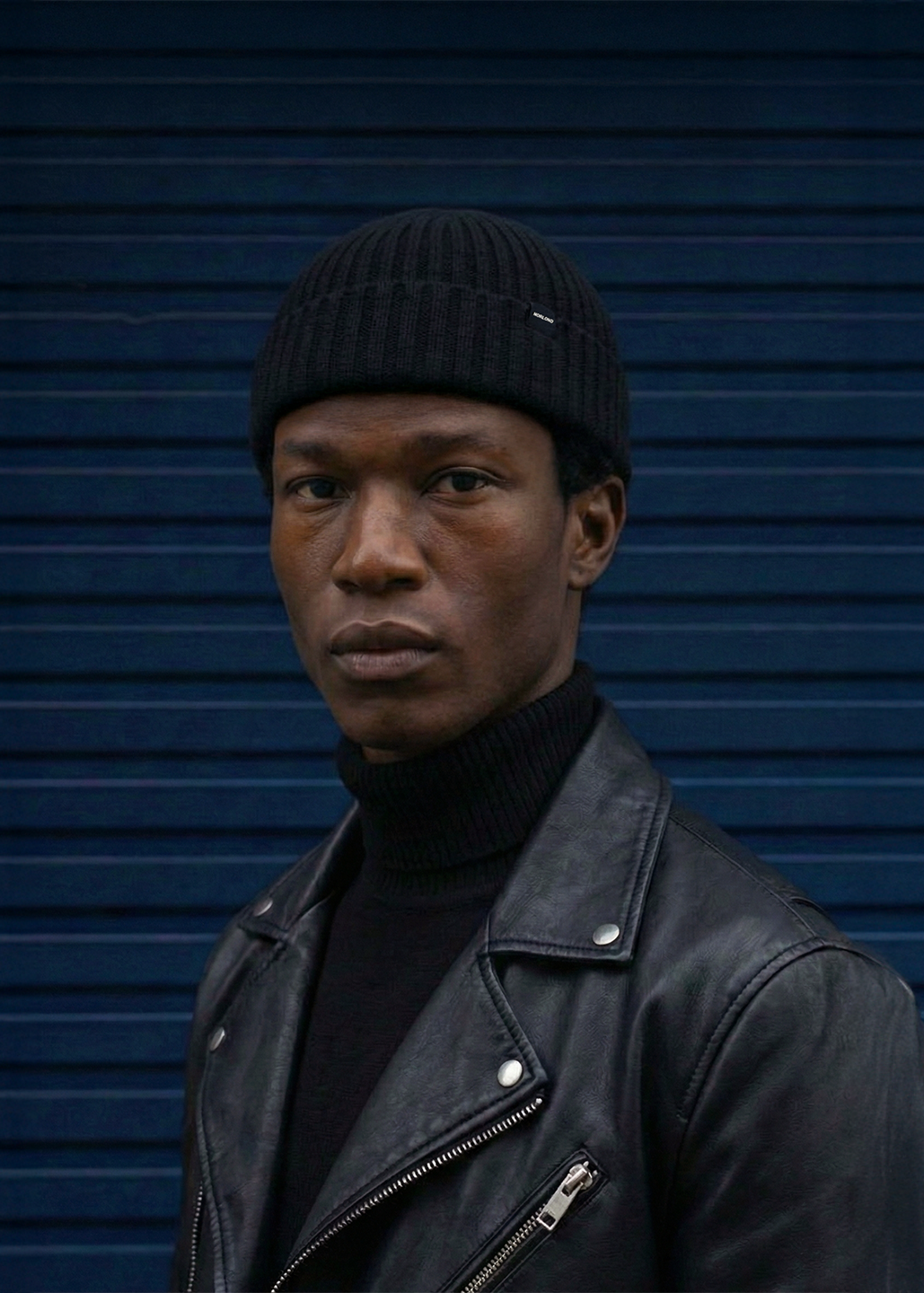 Man wearing a black leather jacket and Black fisherman beanie against a blue background