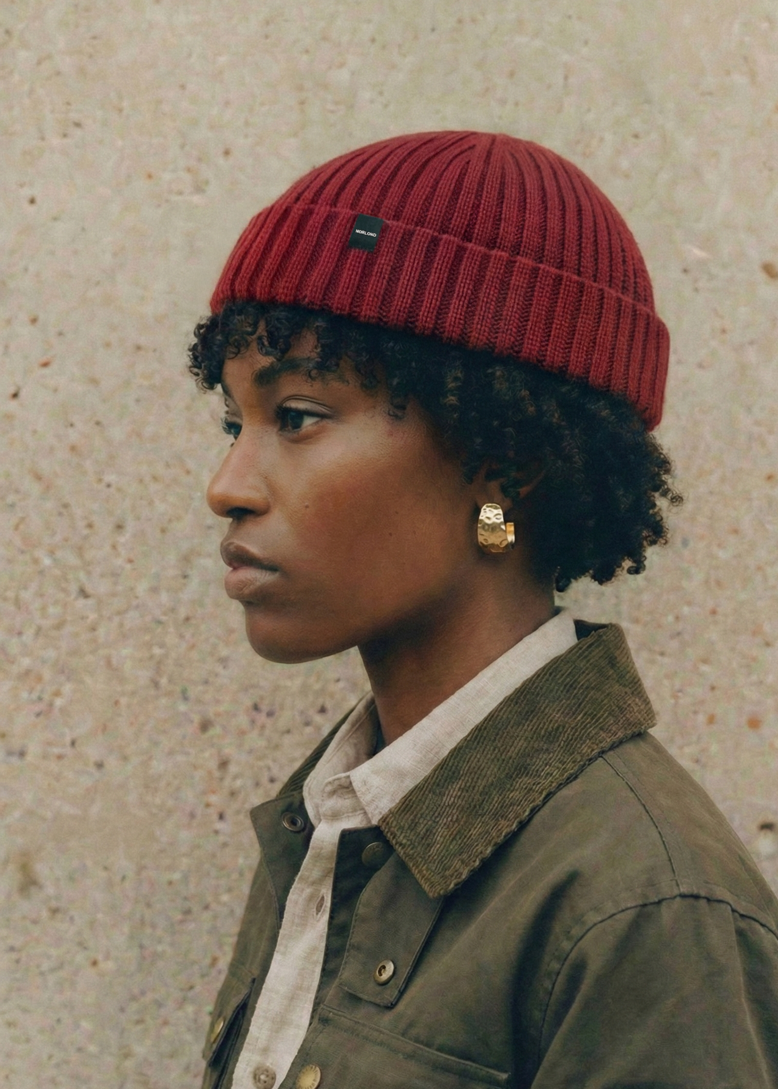 Person wearing a red Fisherman beanie and green jacket against a beige wall