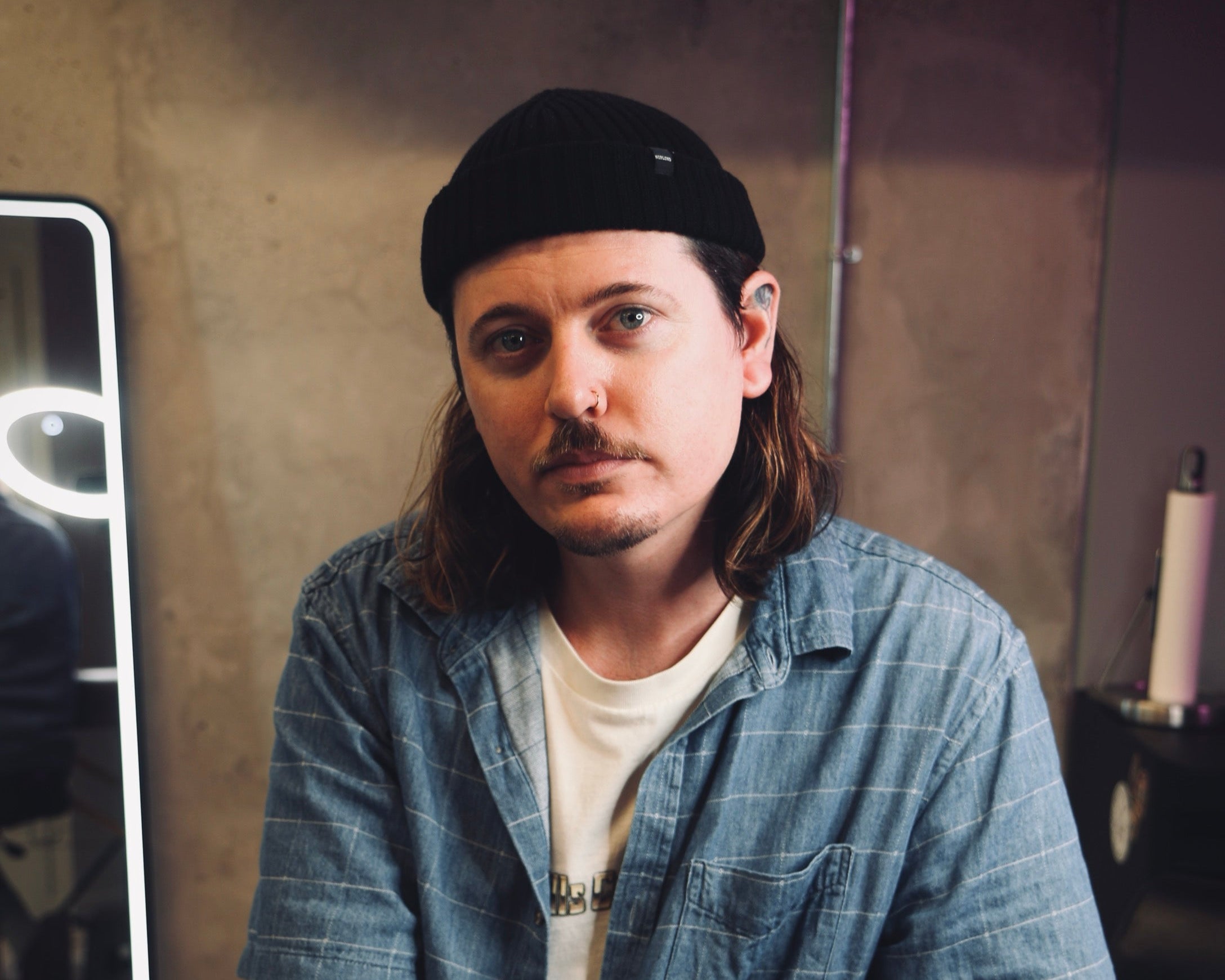 Man with tattoos wearing a denim shirt and black fisherman beanie sitting on a couch.